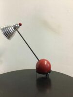 Seitliche Ansicht einer Tischlampe mit futuristischem Design. Die Lampe hat eine kugelförmige Basis in Rot und einen schräg gestellten, silbernen Lampenschirm