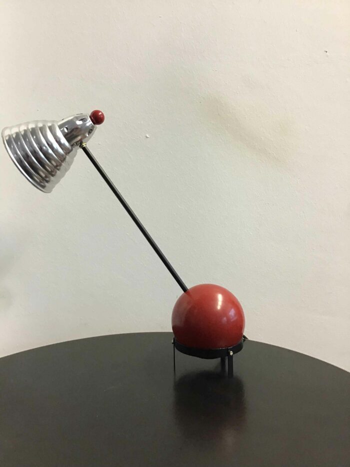 Seitliche Ansicht einer Tischlampe mit futuristischem Design. Die Lampe hat eine kugelförmige Basis in Rot und einen schräg gestellten, silbernen Lampenschirm