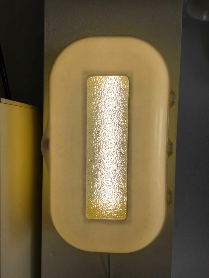 Nahaufnahme einer eingeschalteten Retro-Wandlampe in Gelb mit strukturiertem Glasfenster, das warmes Licht durchlässt