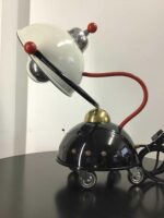 Schreibtischlampe im Ufo-Stil mit schwarzen, glänzenden Rädern und einem flexiblen, roten Arm, der den weißen Lampenschirm hält. Ein ausgefallenes Dekorationsstück für kreative Arbeitsumgebungen.